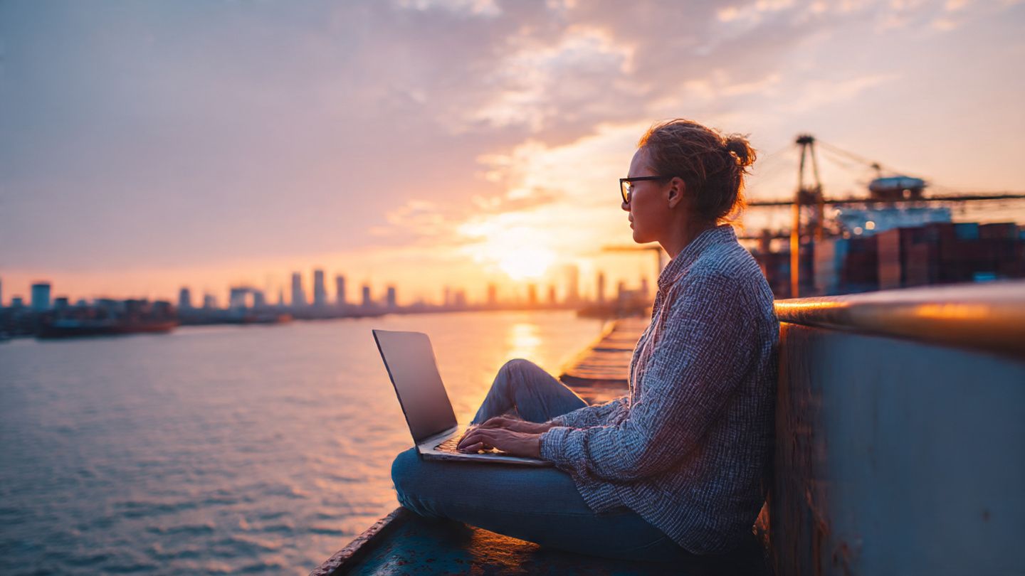 Descriptive image prompt: A business traveler using a laptop on a ship’s deck, with a background of a Southeast Asian port city at sunset, symbolizing modern maritime communication and global business travel