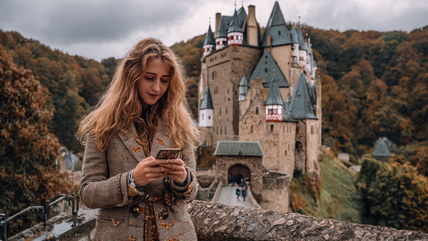 A digital nomad wearing subtle Halloween accessories checks their phone at a European castle during autumn travel, symbolizing seamless roaming across borders