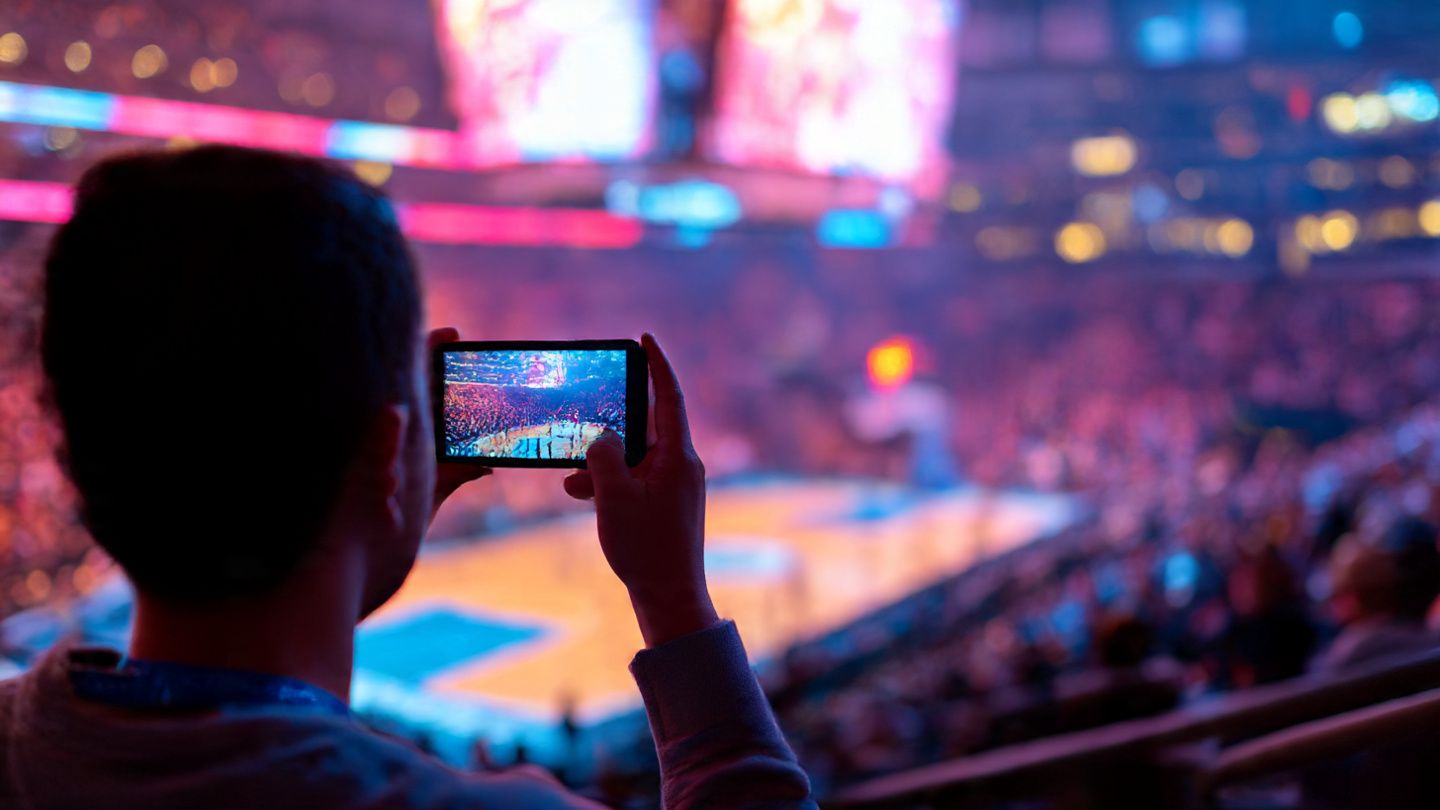 A fan sitting courtside at an NBA arena capturing video on a phone with active 5G signal from an eSIM; vibrant indoor lighting, energetic atmosphere, sports-travel style