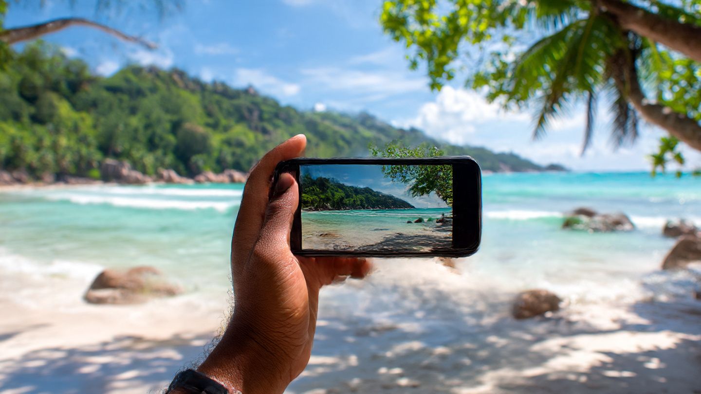 A tourist on Mahé island holding a smartphone against an idyllic Seychelles beach landscape