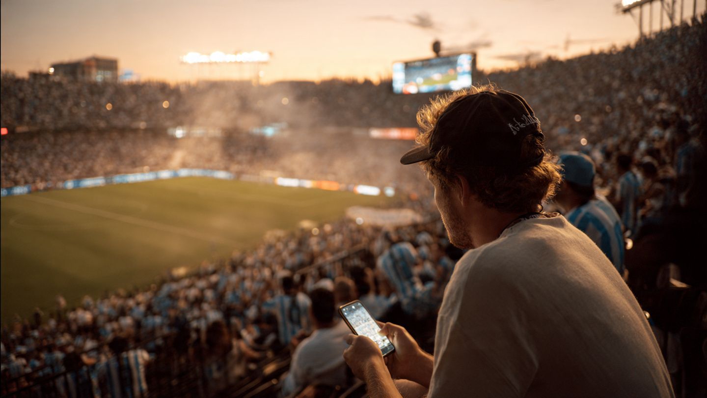 A traveler sitting in GEODIS Park stands before kickoff, phone in hand displaying an active USA eSIM, Messi fans surrounding the area; warm, documentary-style sports moment