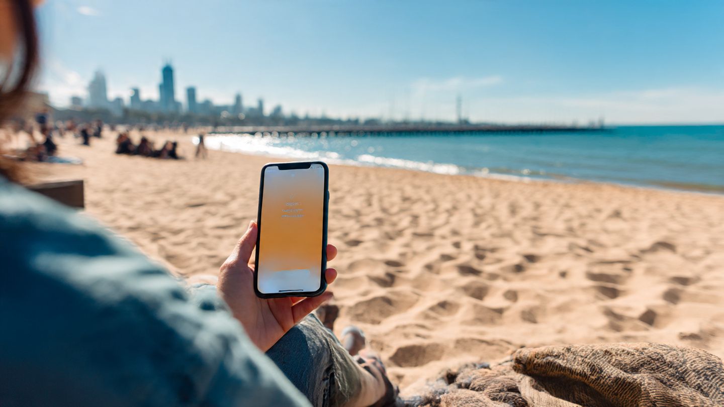 A traveler sitting on Saint Kilda Beach with a phone displaying an eSIM activation screen; bright, modern, adventure-travel photography style