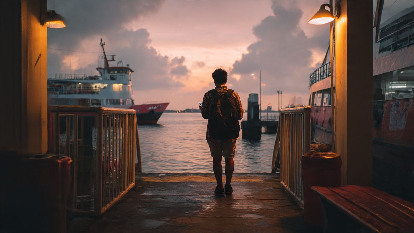 A traveler stepping onto the pier at Batam Centre Ferry Terminal with a phone in hand, warm tropical lighting, cinematic travel aesthetic