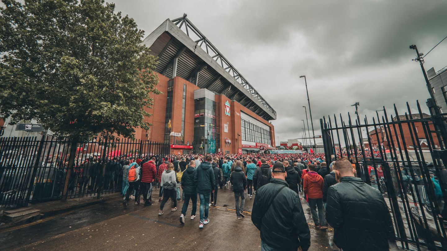 A wide-angle view of Anfield Stadium on matchday, fans entering the gates under cloudy UK skies; sports travel photography, vibrant crowd energy
