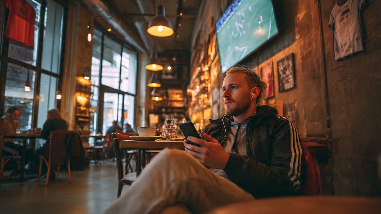 Sports fan using smartphone to watch NFL game highlights in a cozy café overseas, warm evening lighting, relaxed travel lifestyle, digital nomad vibe