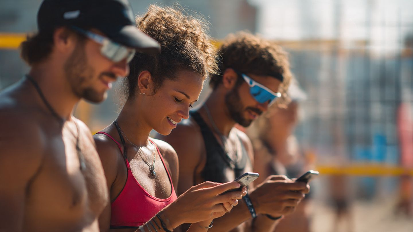 Athletes on a beach volleyball court in Europe checking their smartphones between matches, illustrating eSIM convenience for global sports travel. Warm, lifestyle-focused photo.