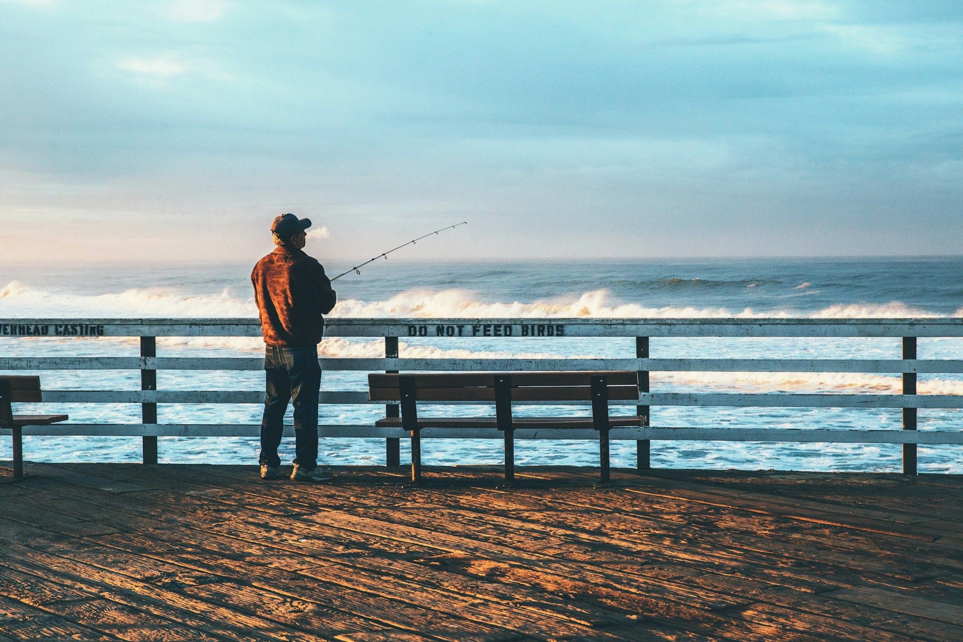 A fisherman casting a line from a wooden pier with waves crashing below.