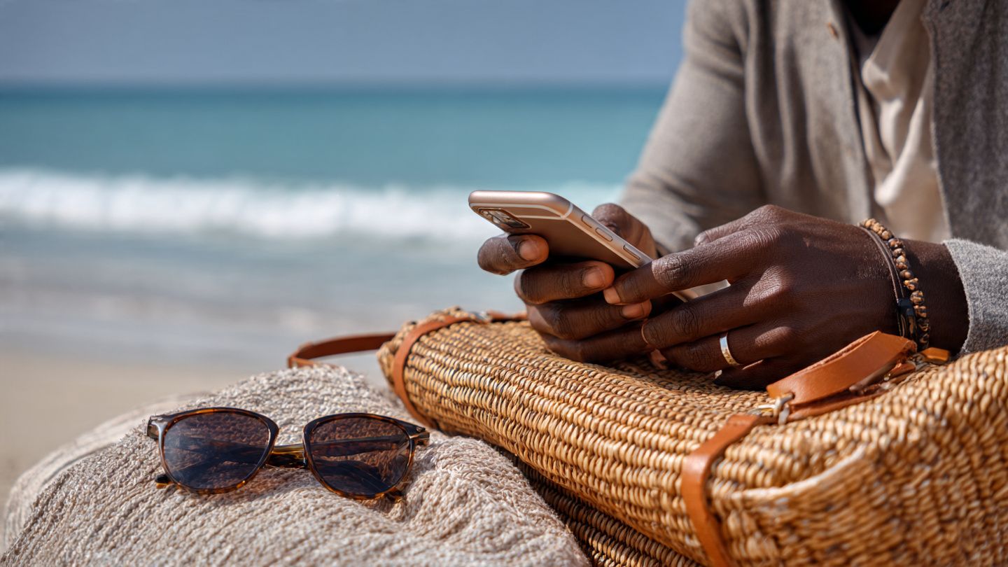 Close-up of a traveler activating an eSIM on a smartphone next to a beach bag and sunglasses on a Jamaican beach; bright daylight, safety and vacation contrast.