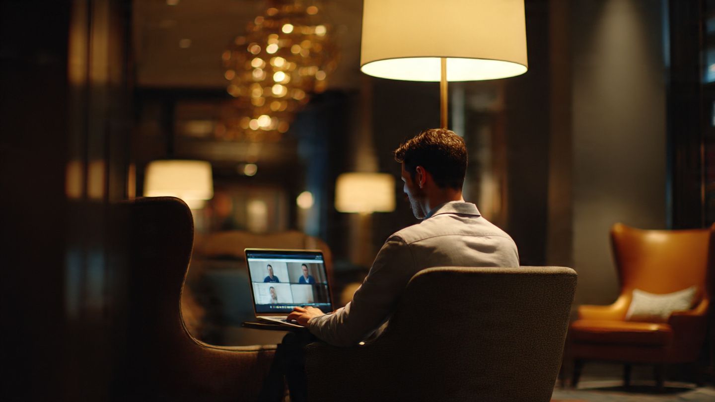 Corporate traveler working from a hotel lounge in New York, laptop open with video conferencing, soft warm lighting, productivity-oriented composition