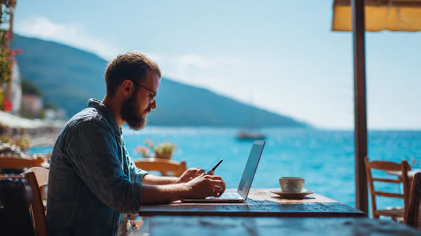Digital nomad in Budva café using laptop and smartphone, showing seamless eSIM connectivity with ocean view background — relaxed workspace, sunny, natural light.