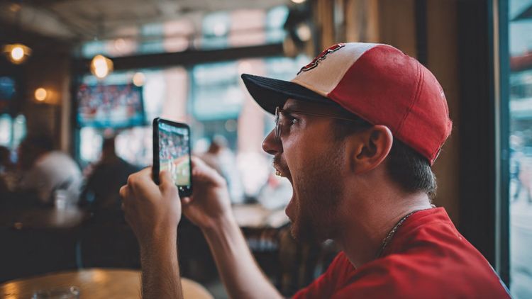 A baseball fan streaming an MLB game on a phone at a European café, showing excitement while wearing a team cap. Bright, dynamic, travel-focused scene