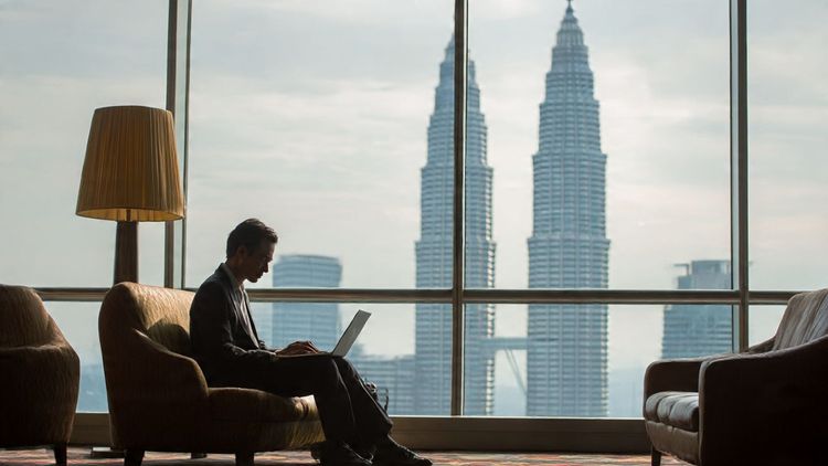 A business traveler in formal attire working on a laptop at an airport lounge in Kuala Lumpur, with the Petronas Towers visible through the window, symbolizing connectivity and mobility.