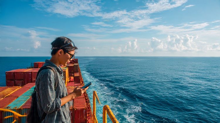 Descriptive image prompt: A cargo ship sailing through the South China Sea under a clear sky, with a professional traveler using a smartphone on deck, representing maritime connectivity and business communication in Southeast Asia