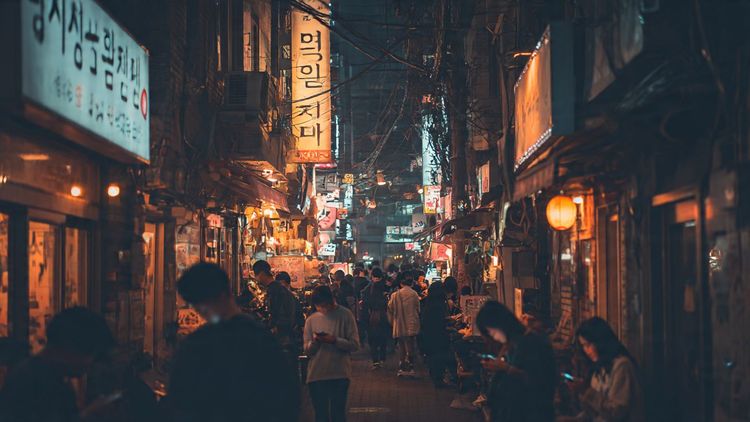 A cinematic street-level view of nighttime Itaewon with neon signs, crowded alleys, and travelers using their phones; warm editorial travel photography style