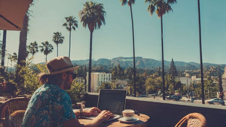 A digital nomad working on a laptop at a sunny Los Angeles café, palm trees and the Hollywood Hills in the background – lifestyle, travel, and tech tone