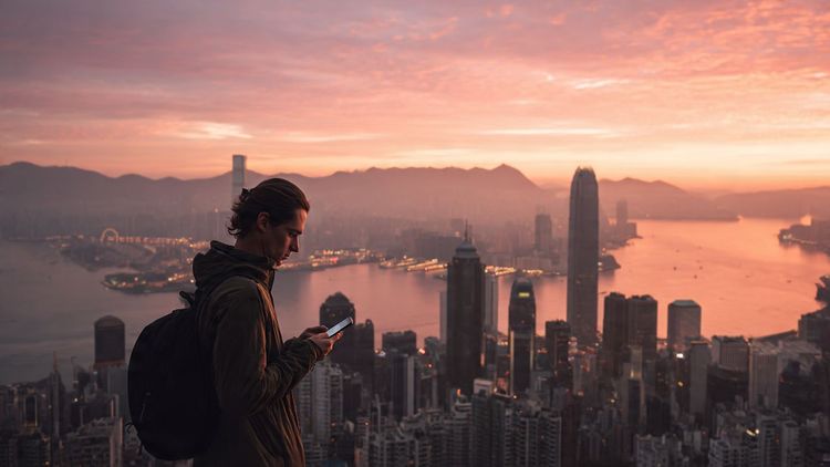 A traveler overlooking Hong Kong’s skyline at sunset while checking their phone, cinematic lighting, editorial travel style