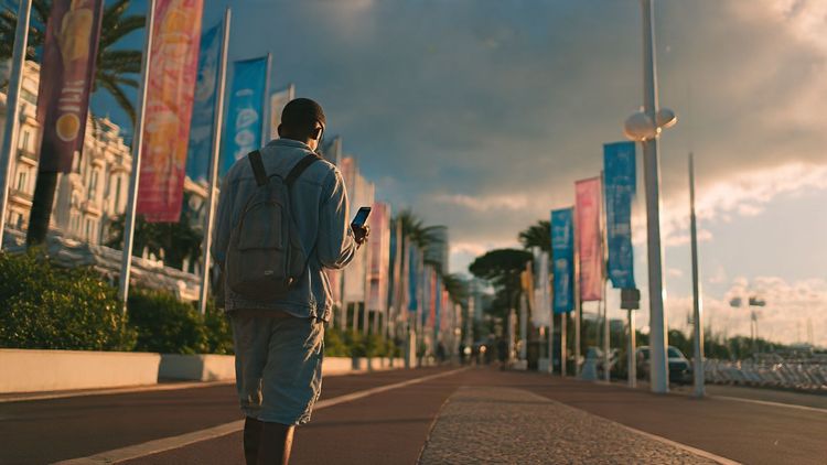A traveler walking along the Croisette in Cannes, holding a phone showing streaming apps while festival banners flutter in the background — cinematic lighting, summer travel mood