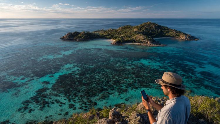 Aerial view of Lizard Island with coral reefs, soft morning light, traveler holding a phone to check signal strength; calm, informative travel-guide tone