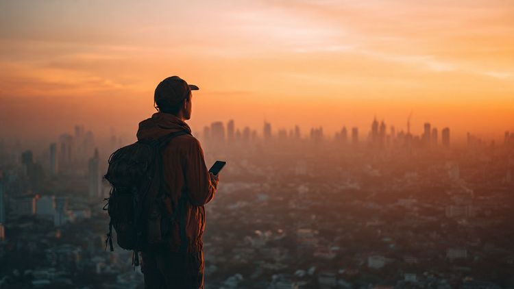 Backpacker standing on a viewpoint overlooking a cityscape in Southeast Asia at sunrise, holding a phone for navigation; warm colors, travel lifestyle, cinematic mood