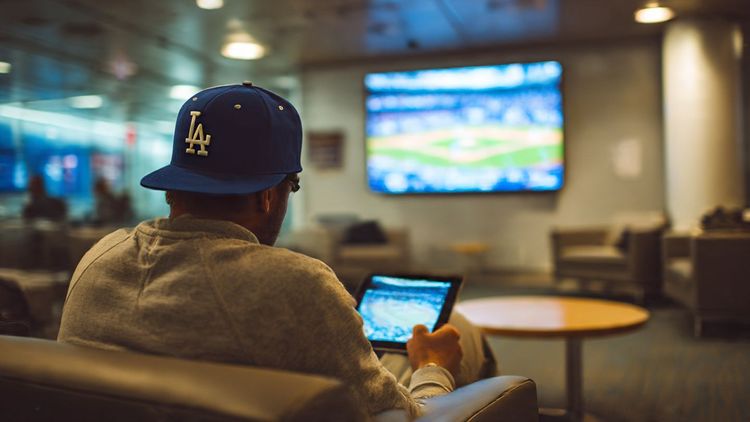 Los Angeles fan in a baseball cap watching a Dodgers game on a tablet while sitting in an airport lounge abroad — modern travel photography with tech and sports vibe