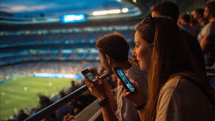 Travelers enjoying a La Liga match at Santiago Bernabéu Stadium in Madrid, sharing highlights on their phones with bright evening lights in the background — travel lifestyle, excitement, sports tourism.