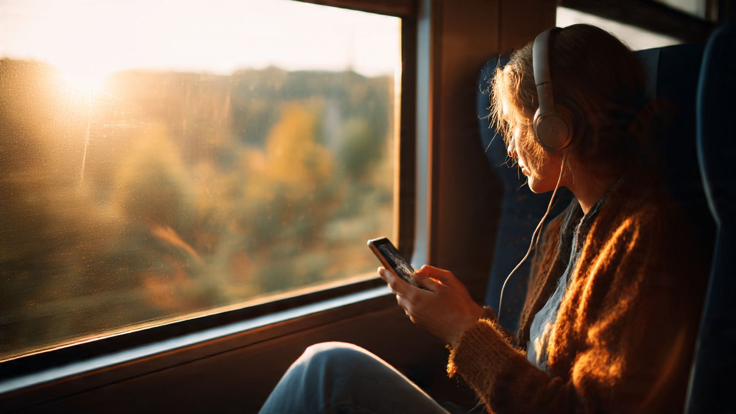 Modern traveler watching a movie on a smartphone on a train, scenic countryside blurred outside, warm natural sunlight, cozy headphones vibe, cinematic travel inspiration