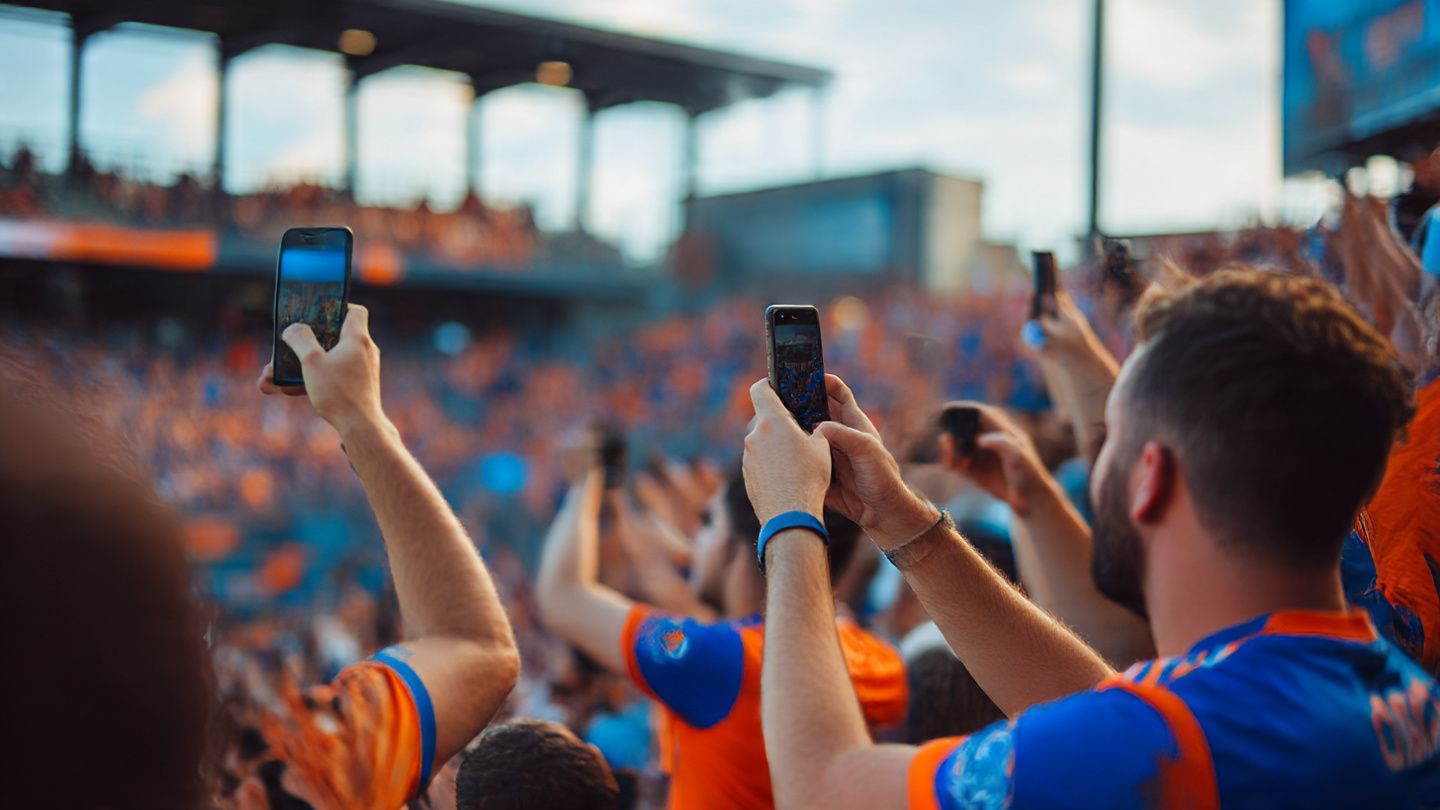 Soccer fans in orange-and-blue FC Cincinnati jerseys using phones in the stadium, sharing photos online, energetic crowd atmosphere