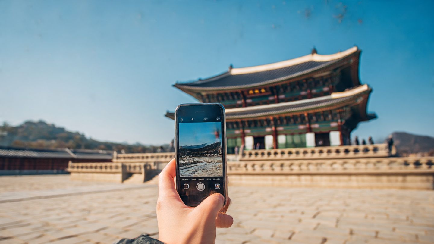 Tourist using smartphone at Gyeongbokgung Palace during daytime, bright blue sky, capturing photos and navigating maps, modern tech meets traditional Korea aesthetic