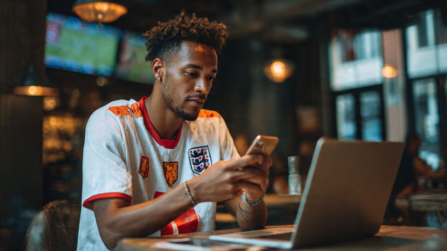 Traveler in sports jersey watching match on phone at European café, laptop open with football stats, crisp travel-sports aesthetic