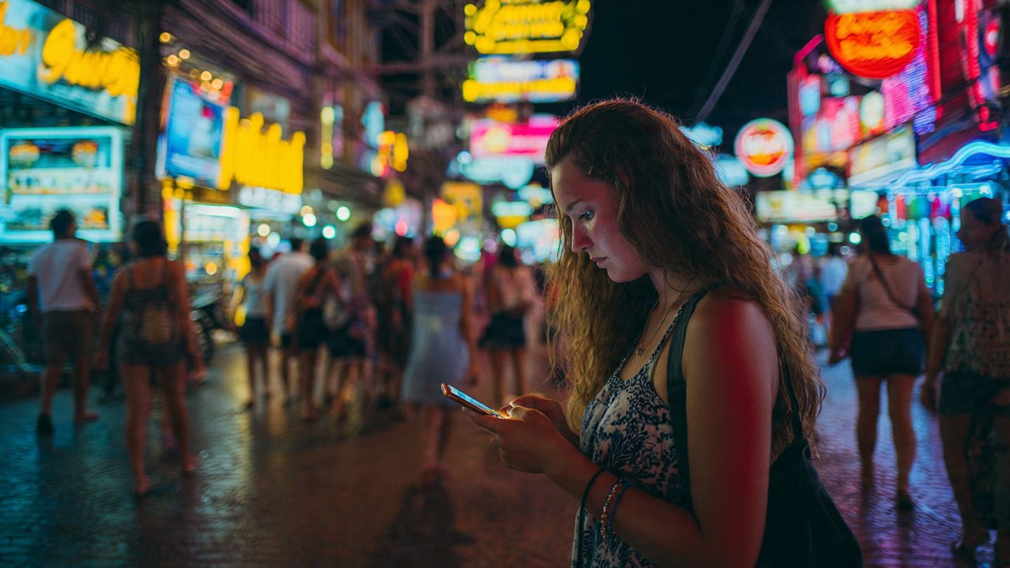 Traveler using a smartphone near Pattaya Walking Street at night, neon lights, busy environment — instructional illustration