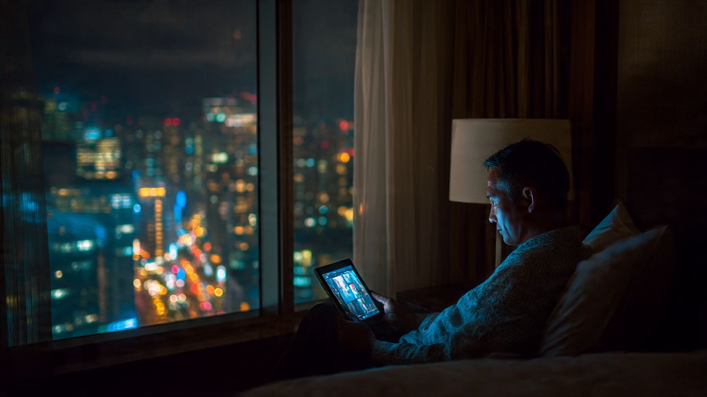 Traveler watching NHK on a tablet in a hotel room at night, city lights outside the window — cinematic photo concept showing stable internet connection abroad