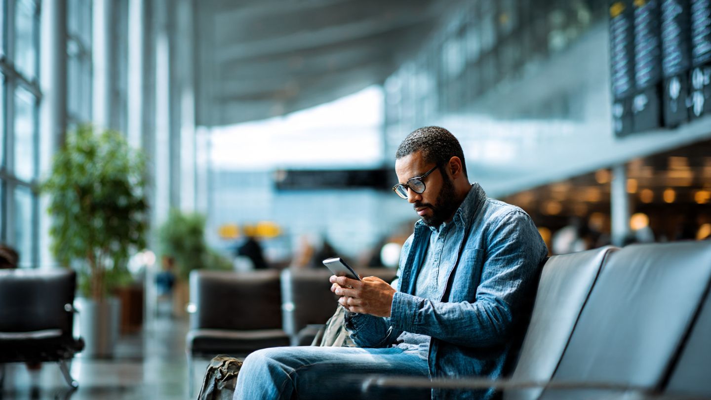 Traveling journalist using phone in airport lounge while scanning global news feeds; modern, bright, informative travel-tech vibe